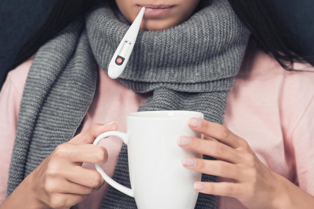plan recadré d'une jeune femme malade mesurant la température avec thermomètre buccal et tenant une tasse de boisson chaude
 - Photo, image