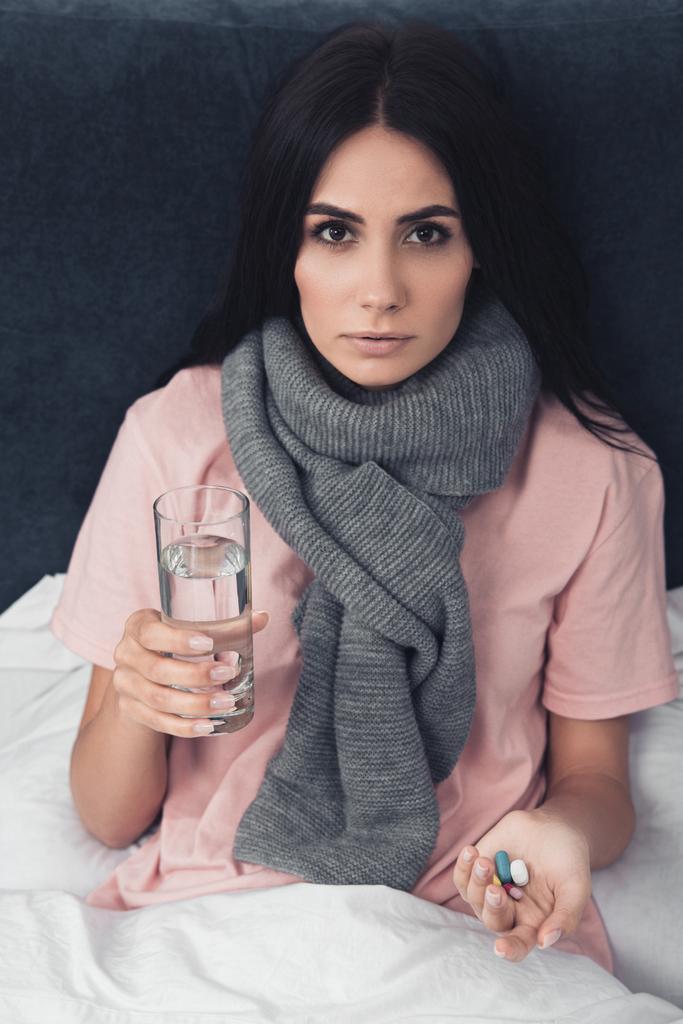 nemocný mladá žena držící pilulky a sklenicí vody když ležíte v posteli - Fotografie, Obrázek