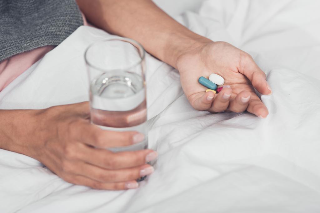 обрезанный снимок больной женщины с таблетками и стаканом воды, лежащей в постели
 - Фото, изображение