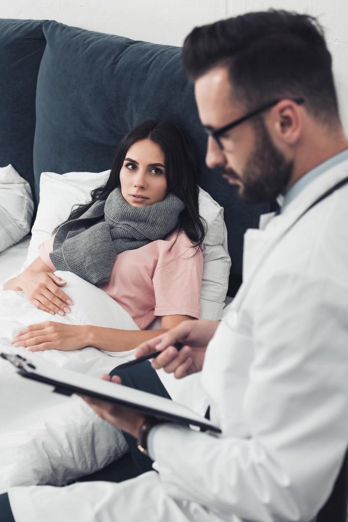 young woman lying in bed while handsome doctor writing in clipboard - Photo, Image