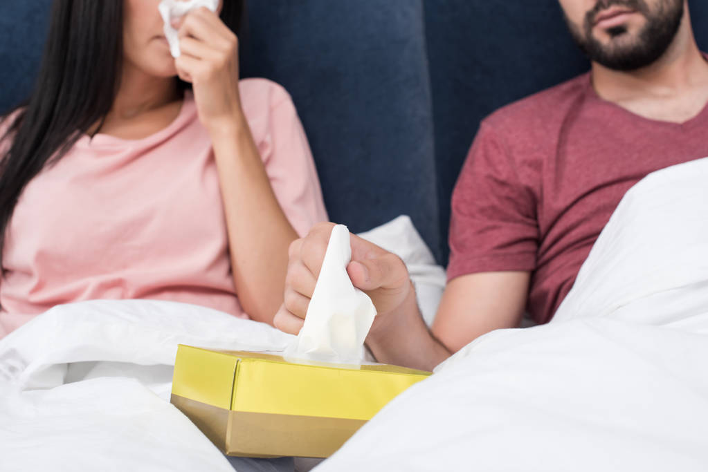 обрезанный снимок больной пары, сидящей в постели и вынимающей бумажные салфетки из коробки
 - Фото, изображение