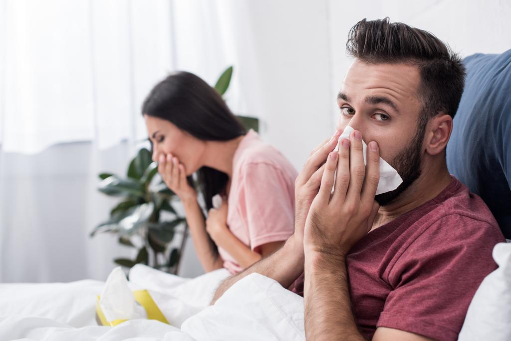 nemocné mladý nemocný pár kýchání do papírové ubrousky při sezení v posteli - Fotografie, Obrázek