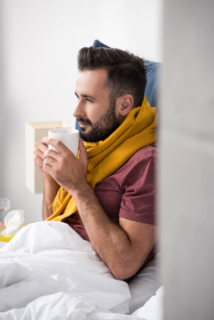 усміхнений хворий молодий чоловік з шарфом тримає чашку гарячого чаю, сидячи в ліжку
 - Фото, зображення