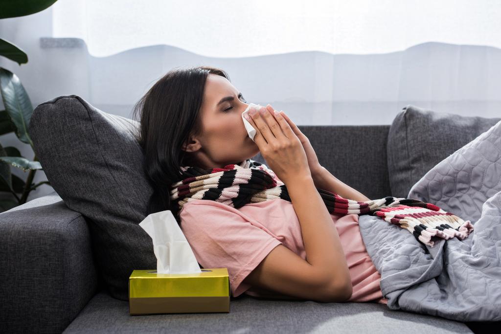 diseased young woman sneezing into paper napkin on sofa - Photo, Image