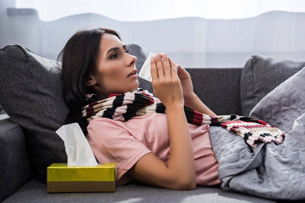 άρρωστος νεαρή γυναίκα φτέρνισμα σε χαρτοπετσέτα στον καναπέ - Φωτογραφία, εικόνα
