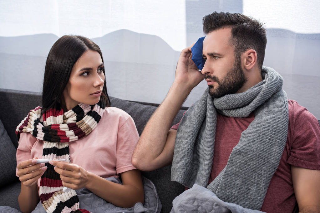 sairas nuori pari, jolla on sähköinen lämpömittari ja jääpakkaus, istuu yhdessä sohvalla
 - Valokuva, kuva