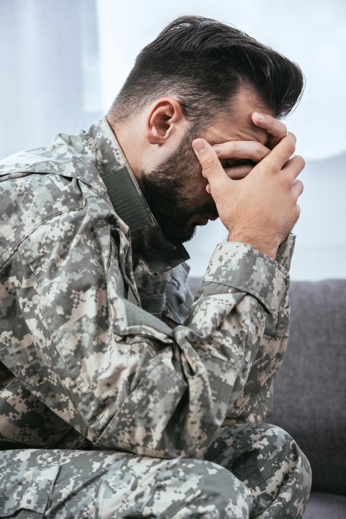 side view of depressed army man in military uniform with post-traumatic stress disorder holding his head while sitting on couch - Photo, Image
