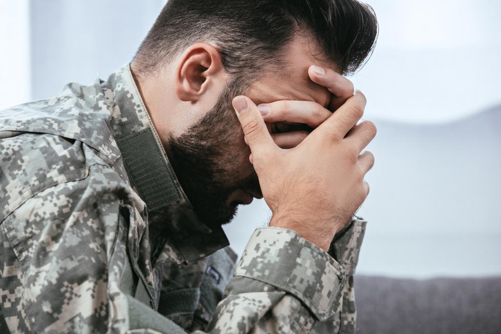 Seitenansicht eines depressiven Armeeangehörigen in Militäruniform mit posttraumatischer Belastungsstörung, der seinen Kopf hält - Foto, Bild