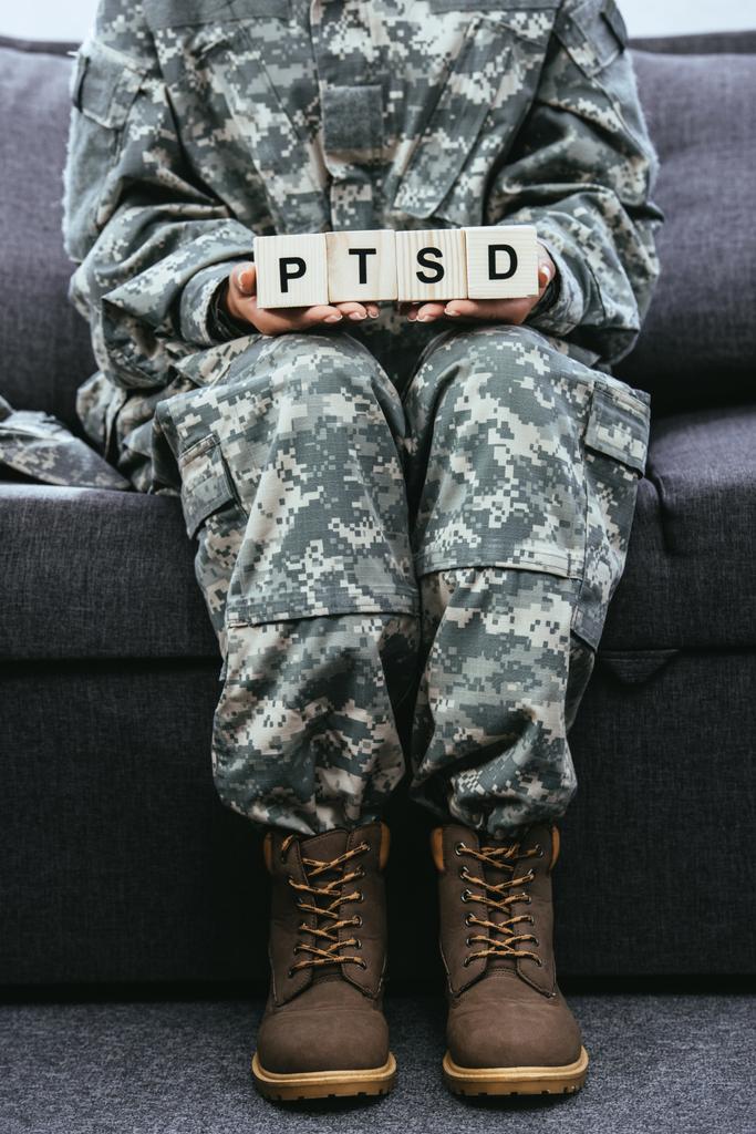 cropped shot of female soldier in military uniform sitting on couch and holding wooden cubes with PTSD sign - Photo, Image