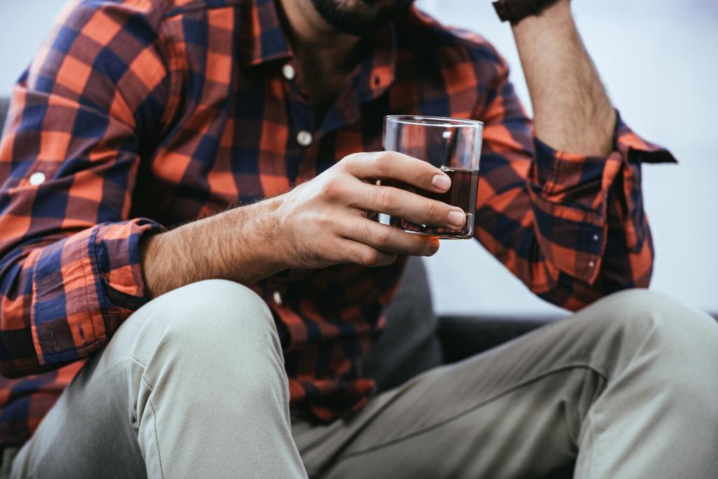 обрезанный снимок молодого человека, держащего стакан виски, сидящего на диване
 - Фото, изображение