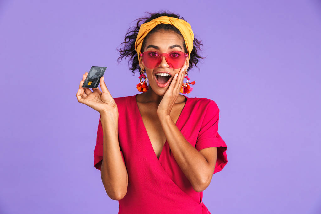 興奮若いアフリカ女性の鉢巻き立っている紫の背景の上にプラスチック製のクレジット カードを示す肖像画 - 写真・画像
