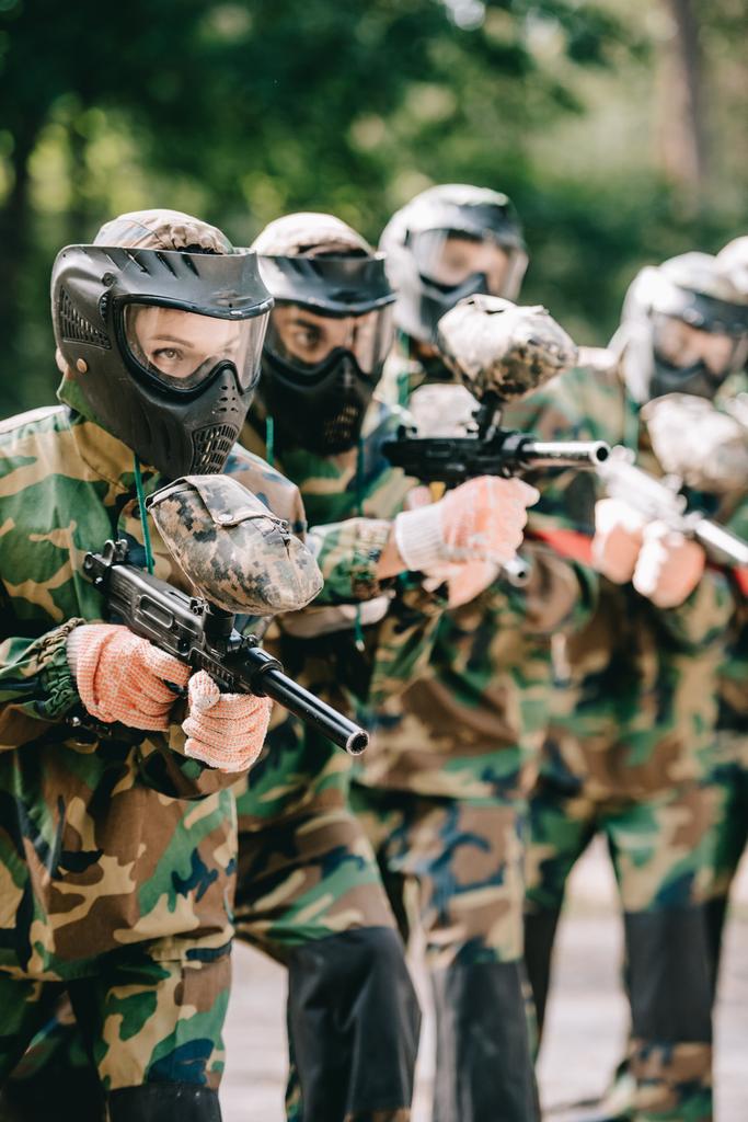 focused female paintball player holding marker gun with her team in protective masks and camouflage playing paintball outdoors  - Photo, Image
