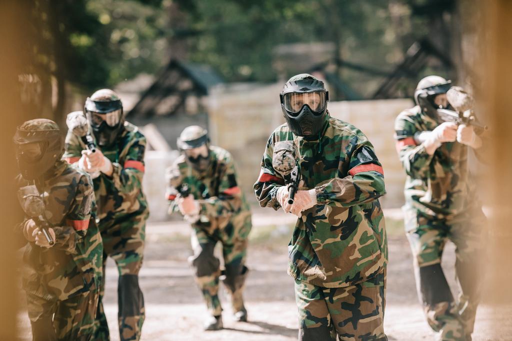 селективный фокус команды по пейнтболу в униформе и защитных масках, играющих в пейнтбол с маркерами на открытом воздухе
 - Фото, изображение