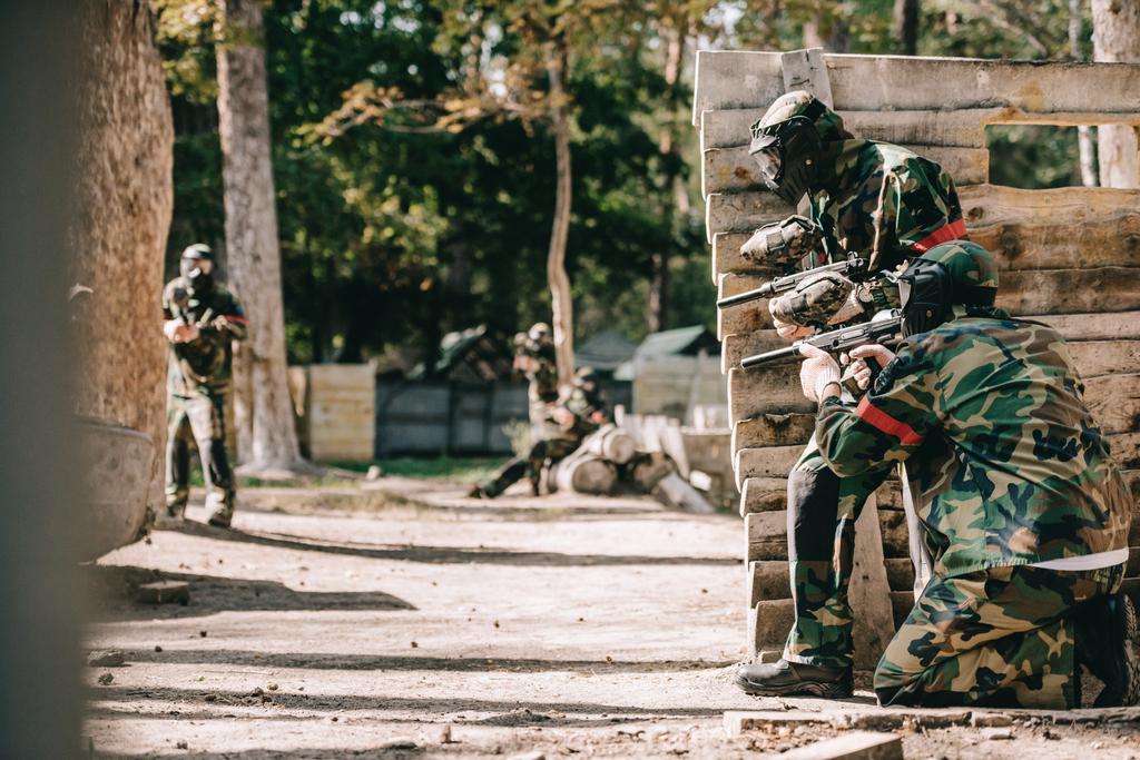 paintball players in camouflage and protective masks aiming with marker guns and hiding behind wooden wall outdoors - Photo, Image