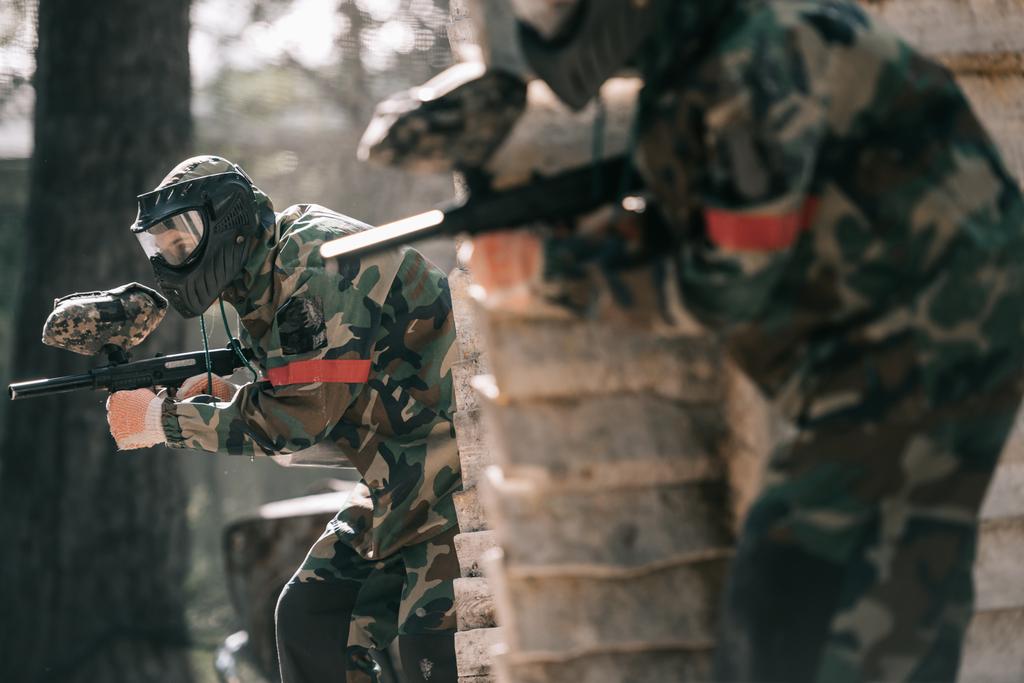 селективный фокус пейнтболиста в защитной маске, держащего маркерную пушку, и его товарища по команде, прячущегося за деревянной стеной
 - Фото, изображение