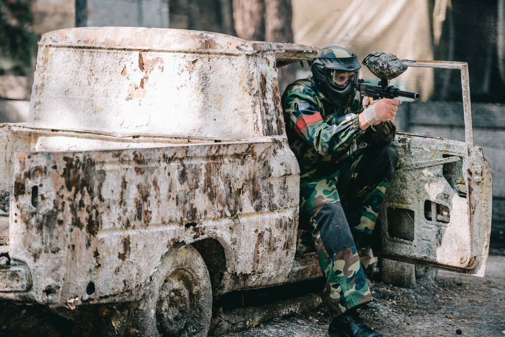 mannelijke paintball speler in goggle masker en camouflage gericht door paintball pistool uit gebroken auto buitenshuis  - Foto, afbeelding