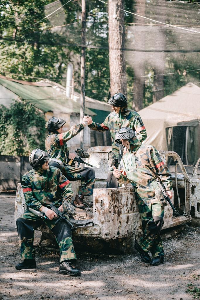 paintball team in camouflage and goggle masks celebrating victory and shaking hands on broken car outdoors  - Photo, Image