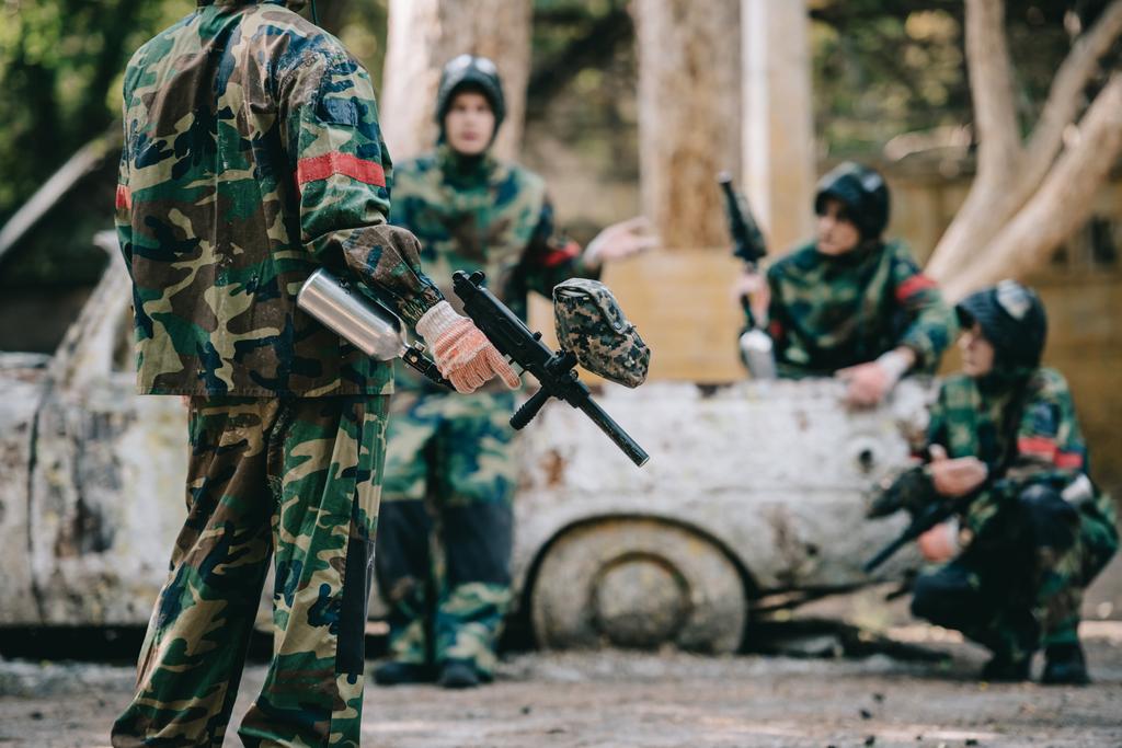 cropped image of paintball team in camouflage uniform with marker guns resting near broken car outdoors  - Photo, Image