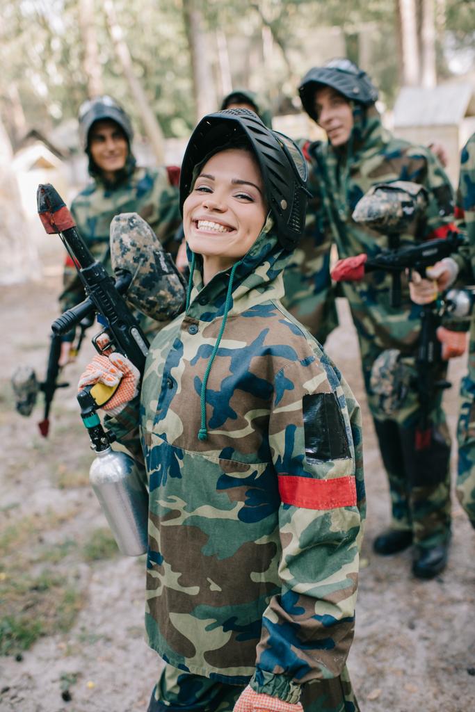 привлекательная улыбающаяся женщина-пейнтболистка в форме держит в руках пейнтбольный пистолет, в то время как ее команда стоит на улице
  - Фото, изображение
