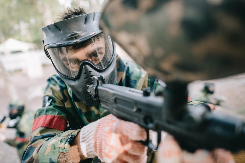 selectieve aandacht voor mannelijke paintball speler in goggle masker en camouflage gericht door paintball pistool buitenshuis  - Foto, afbeelding