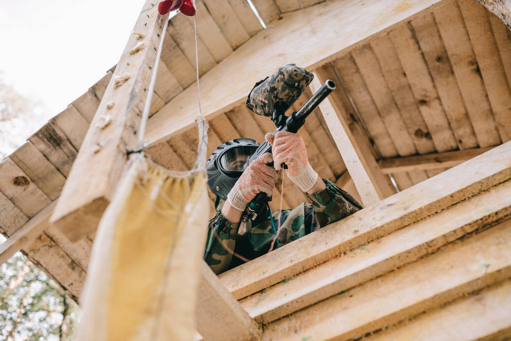 Низкий угол обзора пейнтбольного игрока в очковой маске и камуфляже, прицеливающихся пейнтбольным пистолетом на деревянной башне на открытом воздухе
  - Фото, изображение