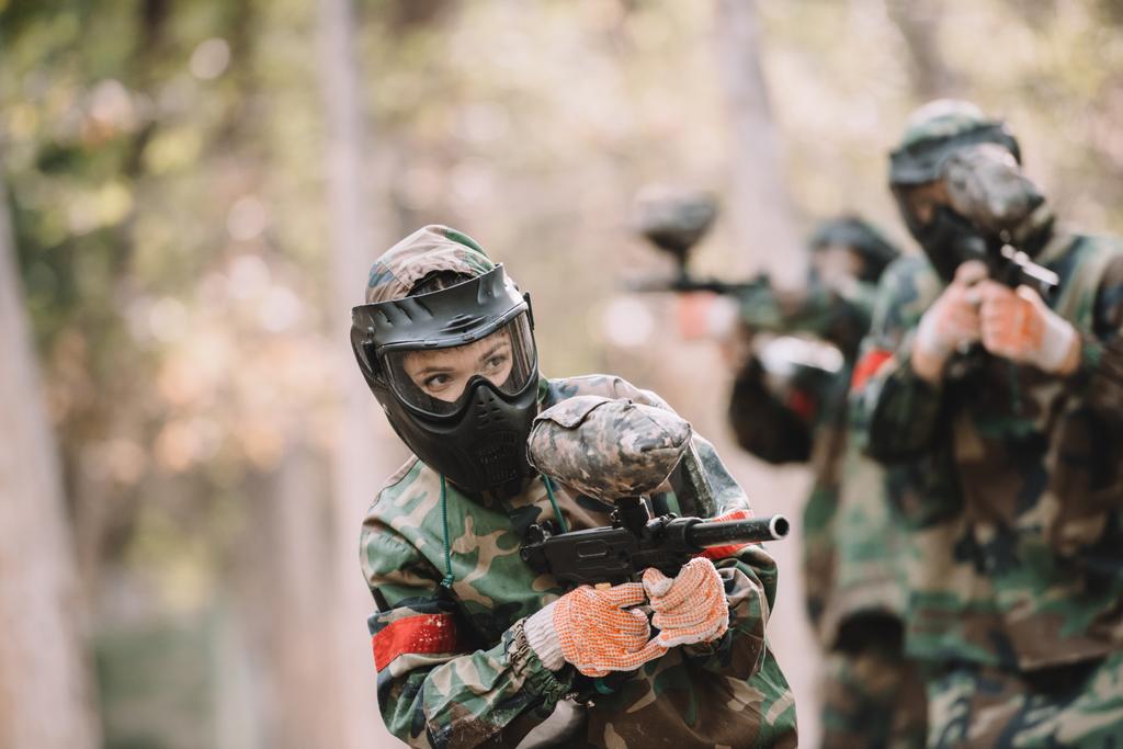 female paintballer in goggle mask and camouflage shooting by marker gun while her team running behind outdoors  - Photo, Image