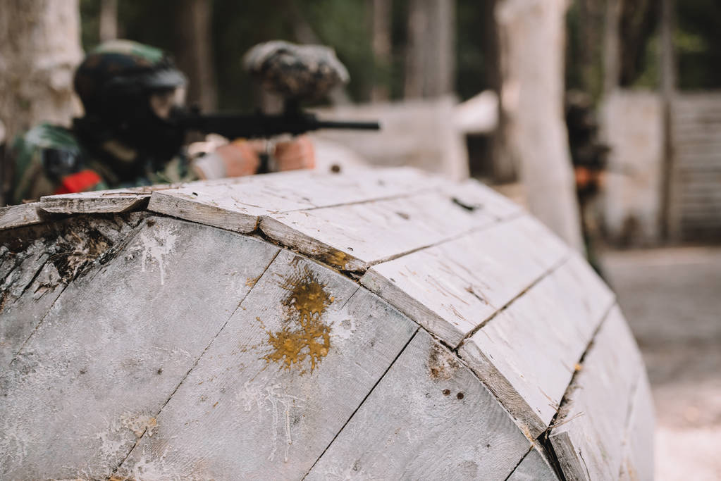 selective focus of paintball splashes on wooden barrel and paintball player shooting behind outdoors  - Photo, Image