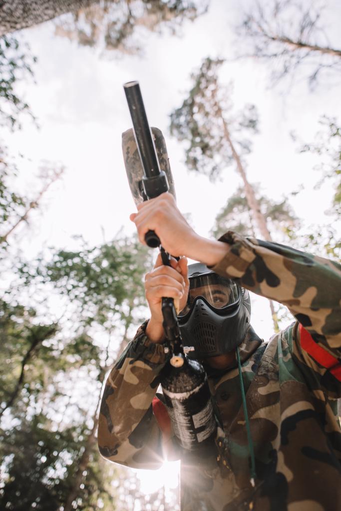 Низкий угол обзора игрока в пейнтбол в очковой маске и камуфляже, прицеливающихся пейнтбольным пистолетом на открытом воздухе
  - Фото, изображение