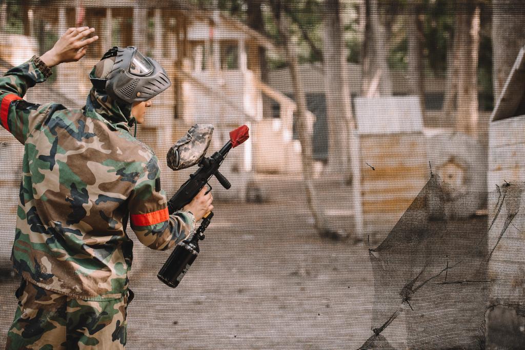 Achteraanzicht van mannelijke paintball speler in goggle masker en camouflage met paintball pistool staande in de buurt van net buiten  - Foto, afbeelding