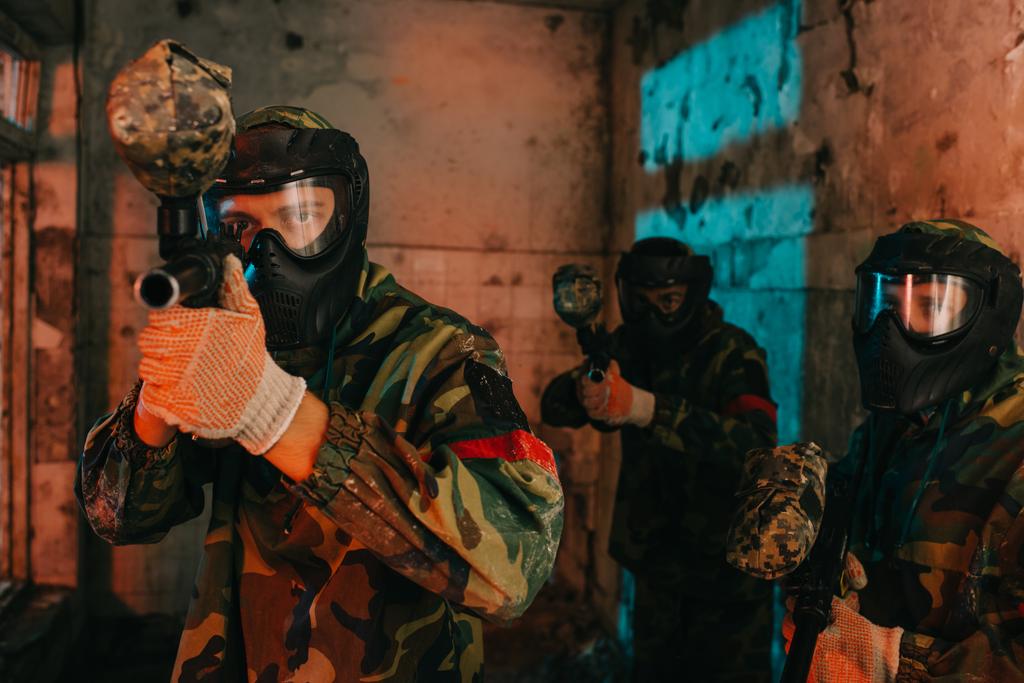 Paintball-Team in Uniform und Schutzmasken spielt Paintball mit Marker-Gewehren in verlassenem Gebäude - Foto, Bild
