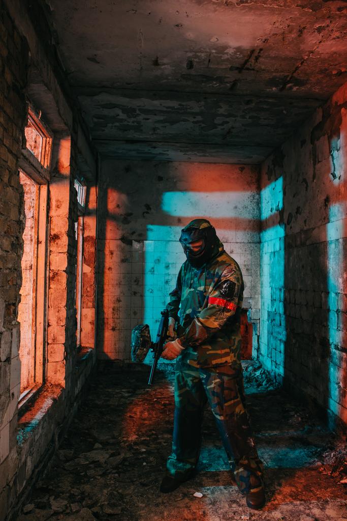 male paintball player in goggle mask and camouflage loading paintball gun in abandoned building - Photo, Image