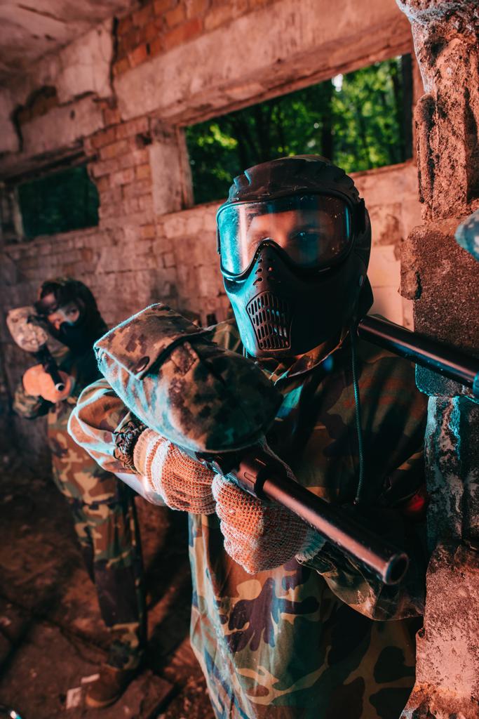 mannelijke paintball speler in goggle masker en uniforme staande terwijl andere speler gericht paintball pistool op zijn nek in verlaten gebouw camouflage  - Foto, afbeelding