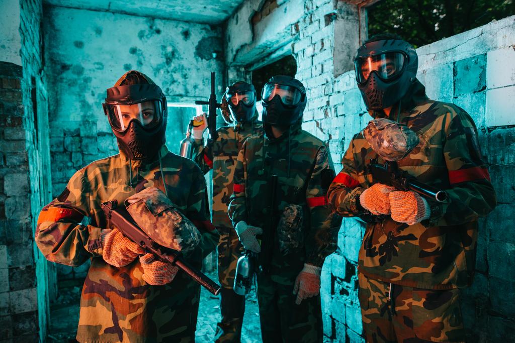 Paintball team in uniform en beschermende maskers permanent met paintball guns in verlaten gebouw - Foto, afbeelding