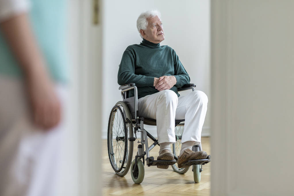 Surullinen vammainen vanhus pyörätuolissa sairaalassa. Hämärtynyt sairaanhoitaja etualalla
 - Valokuva, kuva