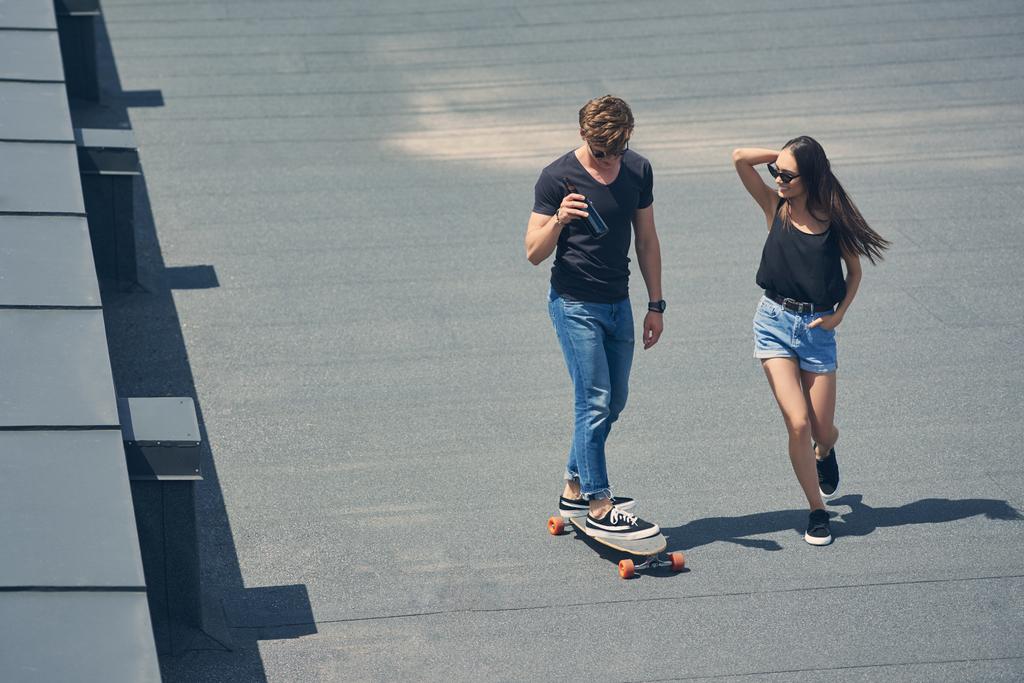 stylish man with bottle of beer skateboarding while asian girlfriend running near - Photo, Image