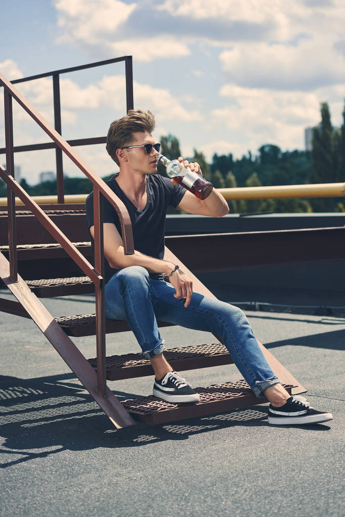 καλαίσθητο άνθρωπος πίνει ρούμι, ενώ κάθεται σε σκαλοπάτια στέγης - Φωτογραφία, εικόνα