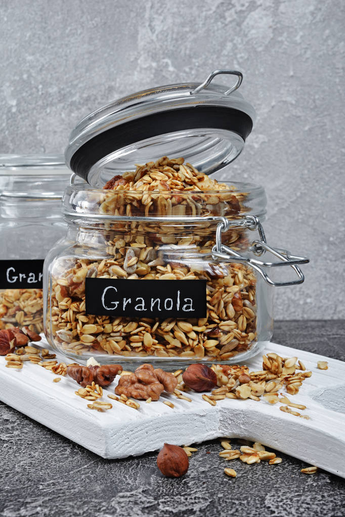 Desayuno saludable granola casera con nueces y semillas de girasol en frascos de vidrio sobre tabla de madera blanca y fondo de hormigón gris
 - Foto, imagen