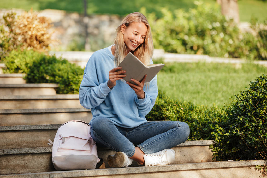 Porträt einer fröhlichen jungen Studentin mit Rucksack, die auf Stufen im Freien sitzt und Buch liest - Foto, Bild