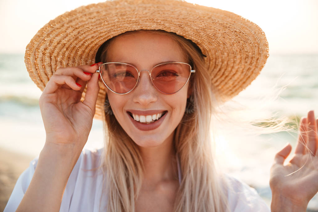Close up de uma menina bonita em chapéu de verão e óculos de sol na praia olhando para a câmera
 - Foto, Imagem