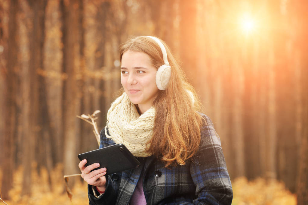 Νεαρό κορίτσι με ζεστό κασκόλ και ωτοασπίδες κρατώντας μαύρα tablet σε Φθινοπωρινό δάσος με θολή φόντο - Φωτογραφία, εικόνα