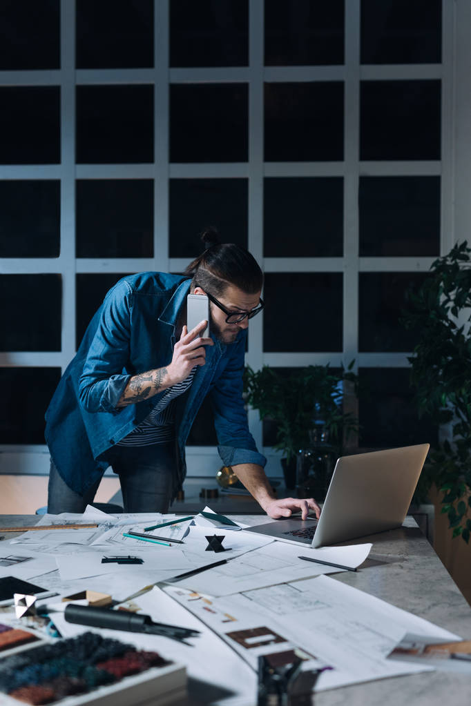 Guapo joven diseñador caucásico hablando en su teléfono móvil mientras trabaja en una oficina por la noche
. - Foto, Imagen
