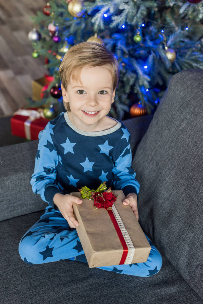 entzückendes glückliches Kind im Pyjama mit Weihnachtsgeschenk und lächelnd in die Kamera - Foto, Bild