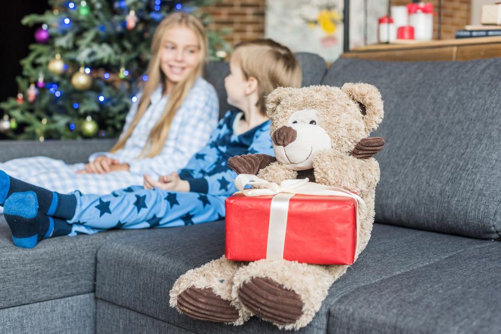 γκρο πλαν θέα αρκουδάκι με δώρο Χριστουγέννων και τα παιδιά στο πιτζάμες που κάθεται στον καναπέ, πίσω από - Φωτογραφία, εικόνα