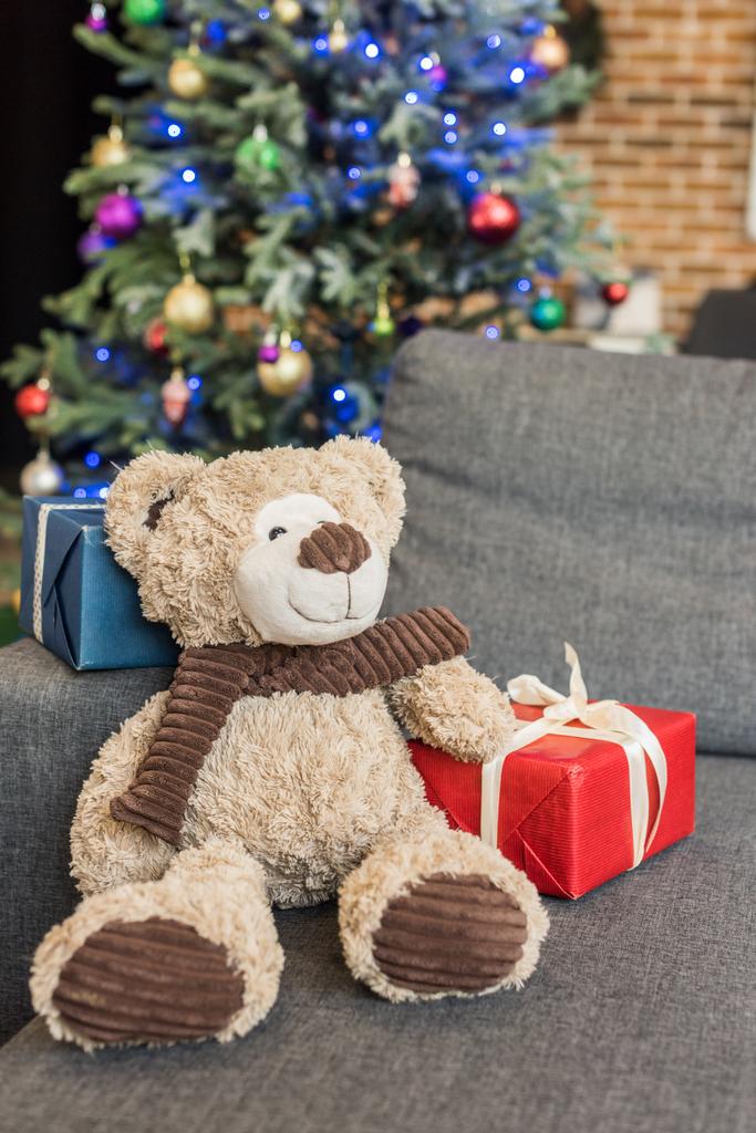 vergrote weergave van teddybeer met geschenkdozen op de sofa en versierde kerstboom achter    - Foto, afbeelding