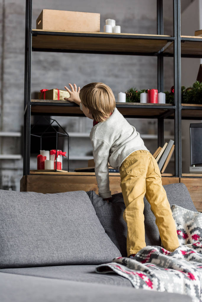 visão traseira da criança de pé no sofá e alcançar o presente de Natal
 - Foto, Imagem