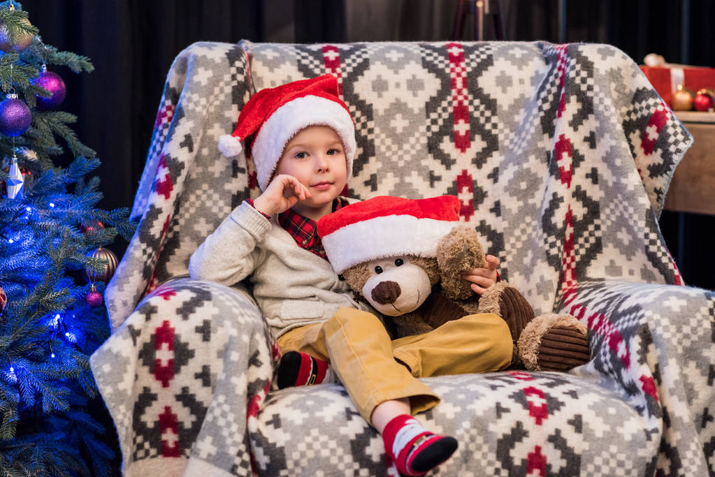 очаровательный маленький мальчик держит плюшевого мишку в шляпе Санты и улыбается в камеру на Рождество
  - Фото, изображение