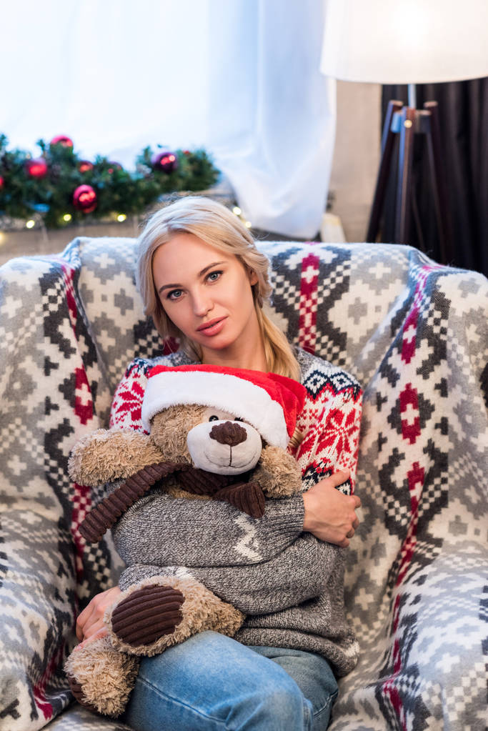 schöne junge Frau umarmt Teddybär in Weihnachtsmütze und schaut in die Kamera - Foto, Bild