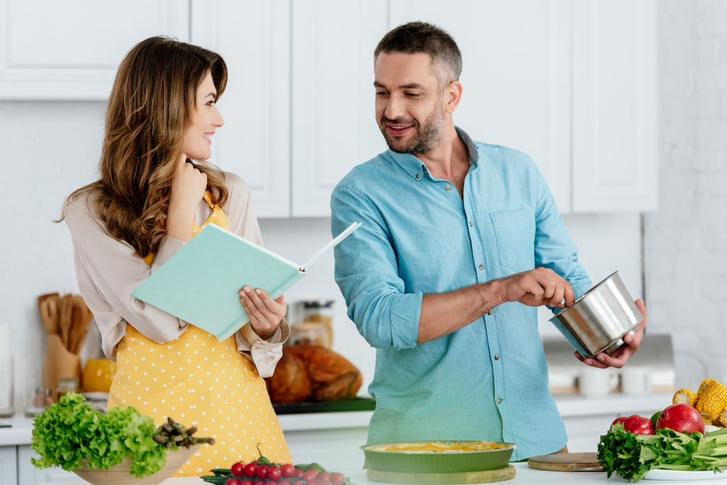 smiling couple reading recipe book while cooking together at kitchen - Photo, Image
