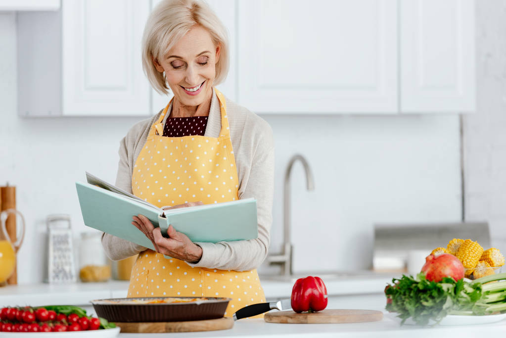 smiling senior woman reading recipe book while cooking at kitchen - Photo, Image
