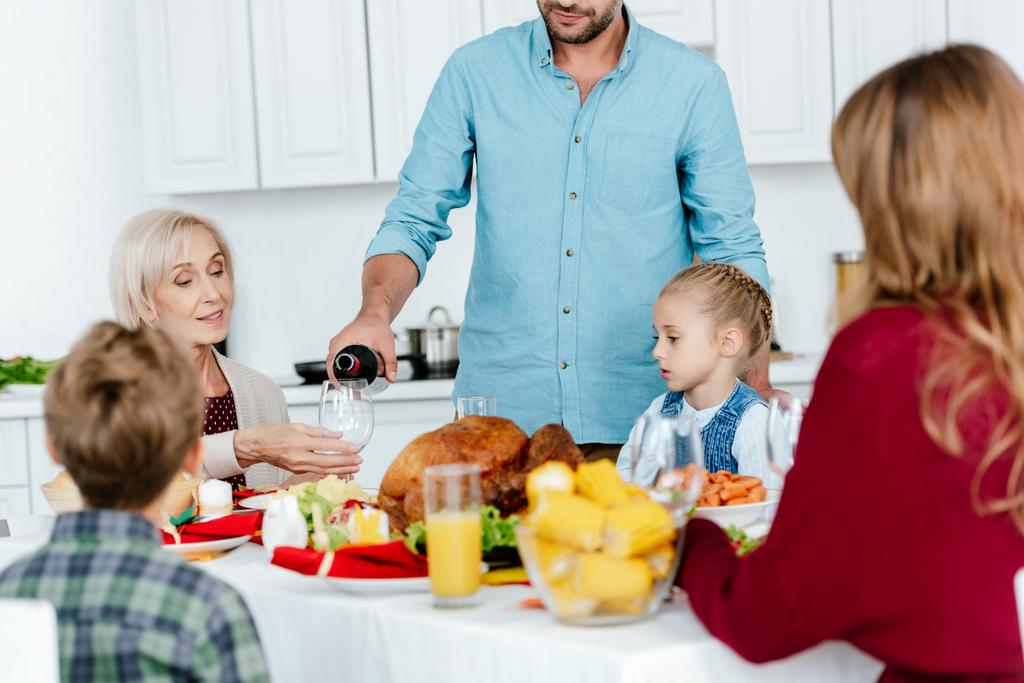 частичный вид человека, наливающего вино в бокал, в то время как семья празднует День благодарения за столом с запечённой индейкой
  - Фото, изображение